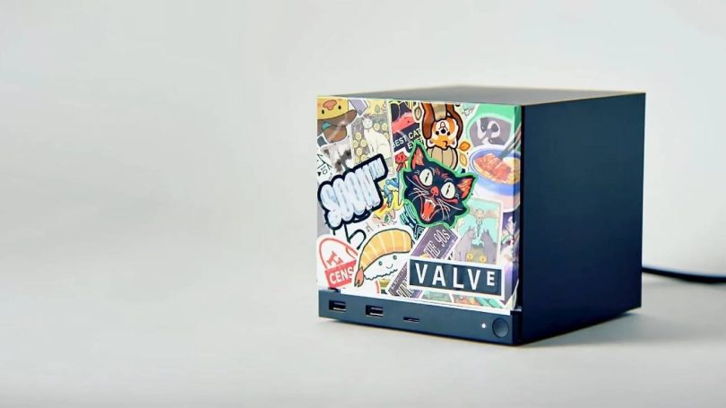 Mini PC Valve Steam Machine noir de forme cubique, posé de trois-quarts face, dont la façade avant est entièrement recouverte de stickers colorés et de graffitis de style pop-culture, incluant un logo officiel "VALVE" en bas à droite et des ports USB en façade.