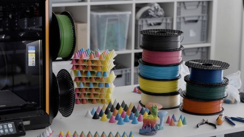 Imprimante 3D sur un plan de travail entourée de bobines de filaments CMYKW et de dizaines de petits cônes de test illustrant les nuances obtenues par la technologie de mélange ColorMix.