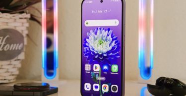 Face avant du smartphone Realme 16 Pro violet, écran OLED allumé affichant l'interface Android avec un fond d'écran de fleur bleue, posé verticalement sur une table blanche entre deux tubes lumineux LED décoratifs.