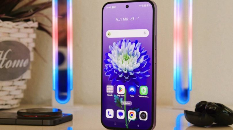 Face avant du smartphone Realme 16 Pro violet, écran OLED allumé affichant l'interface Android avec un fond d'écran de fleur bleue, posé verticalement sur une table blanche entre deux tubes lumineux LED décoratifs.