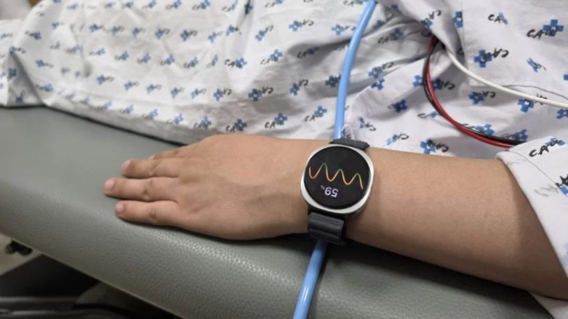 Gros plan sur le poignet d'un patient portant une Samsung Galaxy Watch 6 au bracelet noir en tissu, affichant une courbe de variabilité cardiaque et la valeur 59% lors d'un test médical induit. La montre repose sur la table d'examen grise à côté d'un tube d'eau bleu et sous la blouse d'hôpital à motifs. L'algorithme PPG de la montre analyse les données cardiaques pour prédire les évanouissements 5 minutes à l'avance.