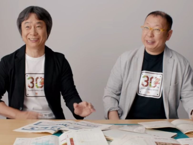 Takashi Tezuka et Shigeru Miyamoto, deux légendes de Nintendo, posent côte à côte avec un t-shirt commémoratif des 30 ans de Super Mario Bros, devant des documents de game design étalés sur une table.