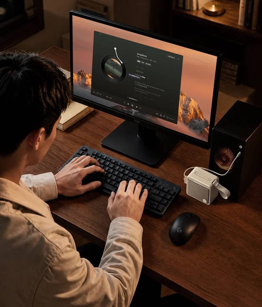 Enceinte Bluetooth Ugreen M3 en version beige posée sur un bureau en bois à côté d'un écran AOC, d'un clavier mécanique et d'une tour PC, illustrant un usage sédentaire en complément d'un setup informatique.