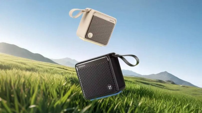Enceinte Bluetooth Ugreen M3 en version noire et beige, avec dragonne et grille acoustique, en lévitation au-dessus d'un paysage de prairie et de montagnes, illustrant son usage outdoor et sa certification IP66.