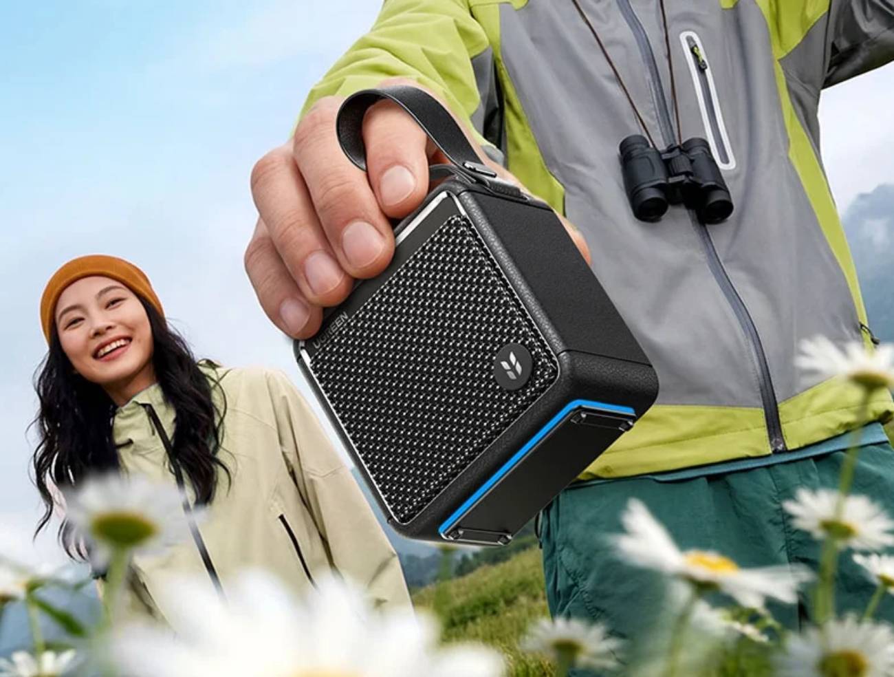 Enceinte Bluetooth Ugreen M3 noire tenue par sa dragonne par un randonneur en veste verte avec jumelles, dans un champ de pâquerettes en montagne, illustrant l'usage outdoor et la portabilité du modèle.