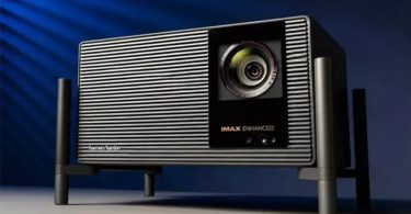 XGIMI X50 Ultra Max vu de 3/4 sur fond bleu studio dramatique, avec son design grille verticale métallique caractéristique, sa lentille principale 70mm centrale, ses badges IMAX Enhanced et harman/kardon, posé sur son piédestal métallique à 4 pieds noirs