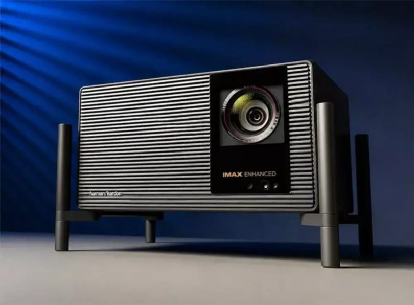 XGIMI X50 Ultra Max vu de 3/4 sur fond bleu studio dramatique, avec son design grille verticale métallique caractéristique, sa lentille principale 70mm centrale, ses badges IMAX Enhanced et harman/kardon, posé sur son piédestal métallique à 4 pieds noirs