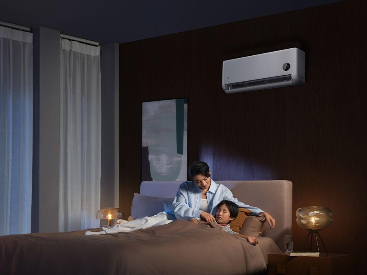 Photographie d'une chambre à coucher à l'ambiance nocturne paisible. Une mère est assise sur un lit et borde délicatement son enfant endormi. Au-dessus d'eux, fixé sur un mur en bois sombre, le climatiseur connecté Xiaomi Mijia fonctionne discrètement. L'image illustre le confort thermique et le fonctionnement silencieux de l'appareil au sein du foyer.