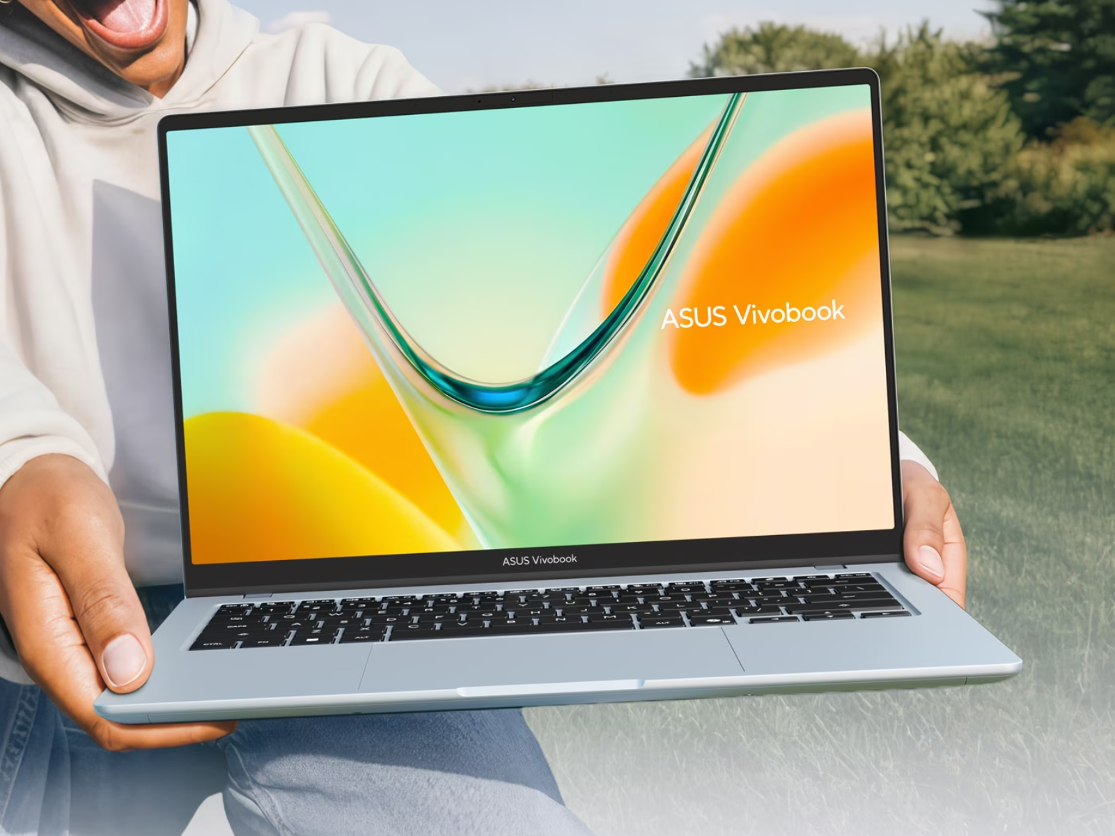 Une personne souriante tenant un ordinateur portable Asus Vivobook S14 bleu clair en extérieur, dans un parc. L'écran OLED très lumineux affiche un fond d'écran coloré, démontrant son excellente lisibilité même en plein jour.