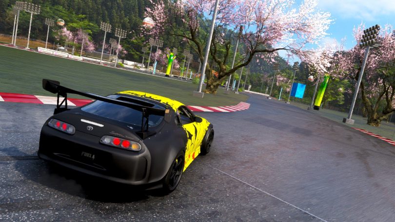 Capture d'écran du jeu Forza Horizon 6 montrant une Toyota Supra noire et jaune négociant un virage sur un circuit asphalté, entouré de cerisiers en fleurs (sakura) typiques des paysages japonais.