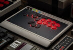 8BitDo Arcade Controller Pro posé sur un bureau, contrôleur arcade sans levier aux boutons rouges rétroéclairés, petit écran intégré et façade noire au cadre gris rétro