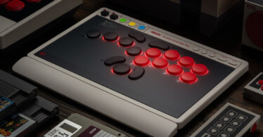 8BitDo Arcade Controller Pro posé sur un bureau, contrôleur arcade sans levier aux boutons rouges rétroéclairés, petit écran intégré et façade noire au cadre gris rétro