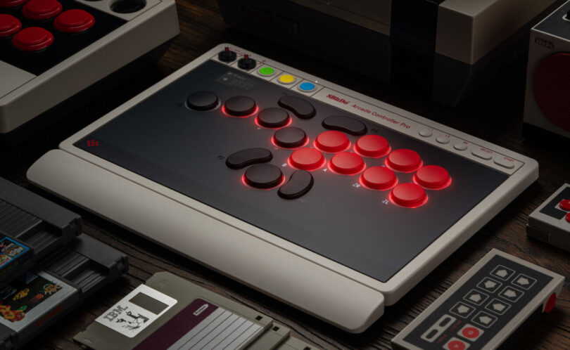 8BitDo Arcade Controller Pro posé sur un bureau, contrôleur arcade sans levier aux boutons rouges rétroéclairés, petit écran intégré et façade noire au cadre gris rétro