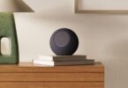 Nouvelle enceinte Amazon Echo Studio avec caméra intégrée, sphère grise anthracite posée sur des livres dans un salon, anneau lumineux bleu allumé