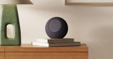 Nouvelle enceinte Amazon Echo Studio avec caméra intégrée, sphère grise anthracite posée sur des livres dans un salon, anneau lumineux bleu allumé