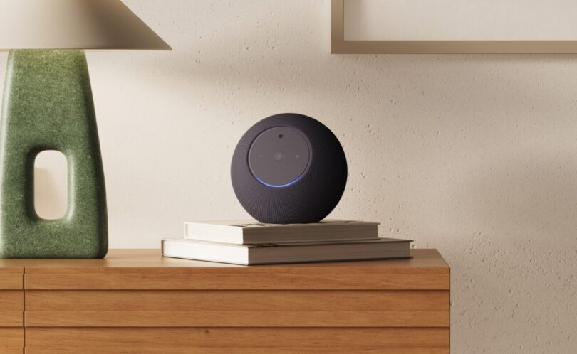 Nouvelle enceinte Amazon Echo Studio avec caméra intégrée, sphère grise anthracite posée sur des livres dans un salon, anneau lumineux bleu allumé