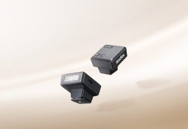 Deux mini flashs Godox iM10 noirs posés sur un fond beige, avec leur tube éclair frontal et leur pied de griffe porte-accessoire