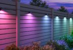Spots Govee Outdoor Deck Lights 2 fixés sur une clôture en bois, éclairant le jardin en rose, violet, bleu et blanc à la tombée de la nuit au-dessus des massifs de fleurs