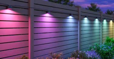Spots Govee Outdoor Deck Lights 2 fixés sur une clôture en bois, éclairant le jardin en rose, violet, bleu et blanc à la tombée de la nuit au-dessus des massifs de fleurs