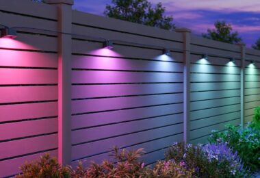 Spots Govee Outdoor Deck Lights 2 fixés sur une clôture en bois, éclairant le jardin en rose, violet, bleu et blanc à la tombée de la nuit au-dessus des massifs de fleurs