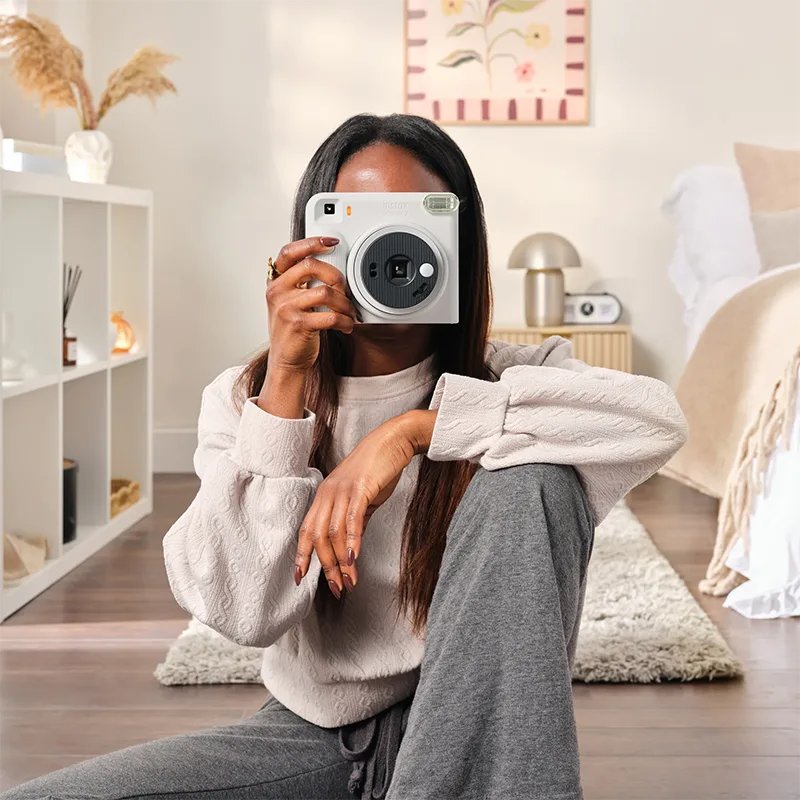 Jeune femme assise dans un salon lumineux qui prend une photo avec un appareil instantané instax SQUARE 2 blanc brumeux tenu devant son visage