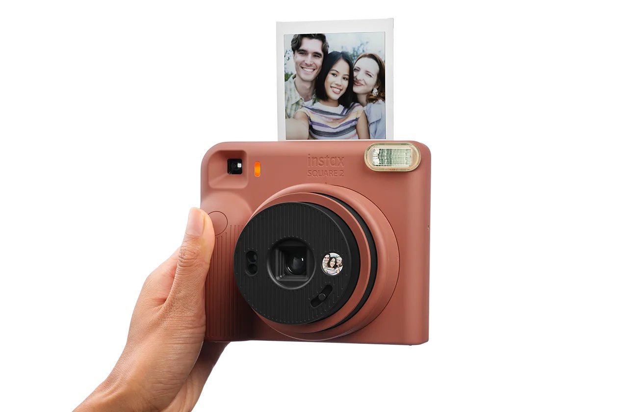 Mains tenant un appareil instax SQUARE 2 rouge brique, objectif tourné vers l'utilisateur, avec un tirage selfie de trois personnes qui sort de l'appareil