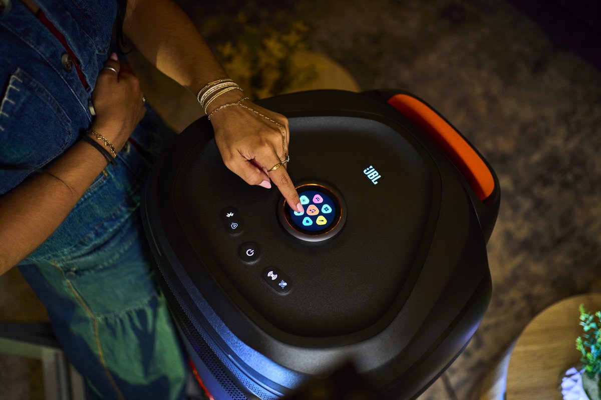 Main appuyant sur l'écran tactile rond aux icônes colorées sur le dessus de la JBL PartyBox Ultimate 2, entouré de boutons physiques et d'une poignée de transport orange