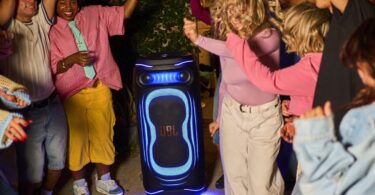 Enceinte de soirée JBL PartyBox illuminée de bleu posée au sol en extérieur de nuit, entourée de jeunes qui dansent, illustrant le lancement des JBL PartyBox Ultimate 2 et Pulse 6