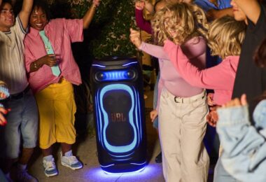 Enceinte de soirée JBL PartyBox illuminée de bleu posée au sol en extérieur de nuit, entourée de jeunes qui dansent, illustrant le lancement des JBL PartyBox Ultimate 2 et Pulse 6