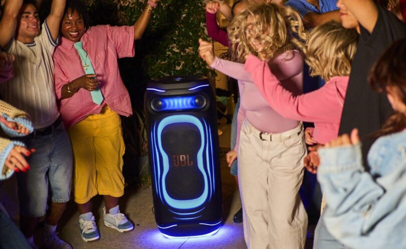 Enceinte de soirée JBL PartyBox illuminée de bleu posée au sol en extérieur de nuit, entourée de jeunes qui dansent, illustrant le lancement des JBL PartyBox Ultimate 2 et Pulse 6