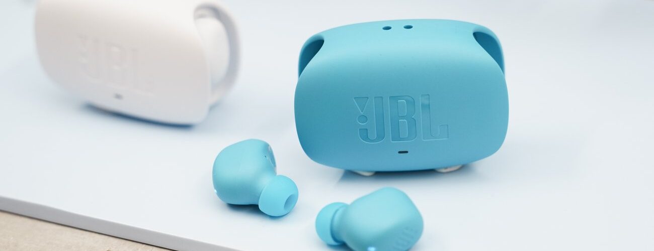 Écouteurs sans fil JBL Wave Buds 3 coloris bleu canard posés devant leur boîtier de charge à double ouverture latérale, avec un second boîtier blanc en arrière-plan
