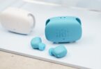 Écouteurs sans fil JBL Wave Buds 3 coloris bleu canard posés devant leur boîtier de charge à double ouverture latérale, avec un second boîtier blanc en arrière-plan
