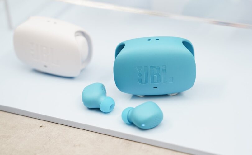 Écouteurs sans fil JBL Wave Buds 3 coloris bleu canard posés devant leur boîtier de charge à double ouverture latérale, avec un second boîtier blanc en arrière-plan