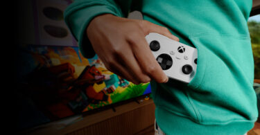Manette XBOX Nano blanche glissée dans la poche d'un sweat vert, devant une TV affichant un jeu, illustrant le format de poche de la nouvelle manette Xbox pour le cloud gaming