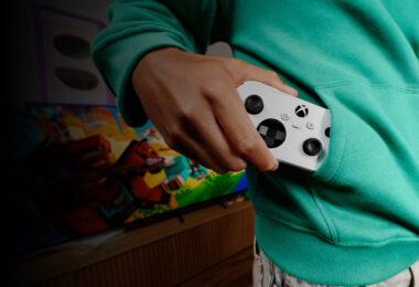 Manette XBOX Nano blanche glissée dans la poche d'un sweat vert, devant une TV affichant un jeu, illustrant le format de poche de la nouvelle manette Xbox pour le cloud gaming