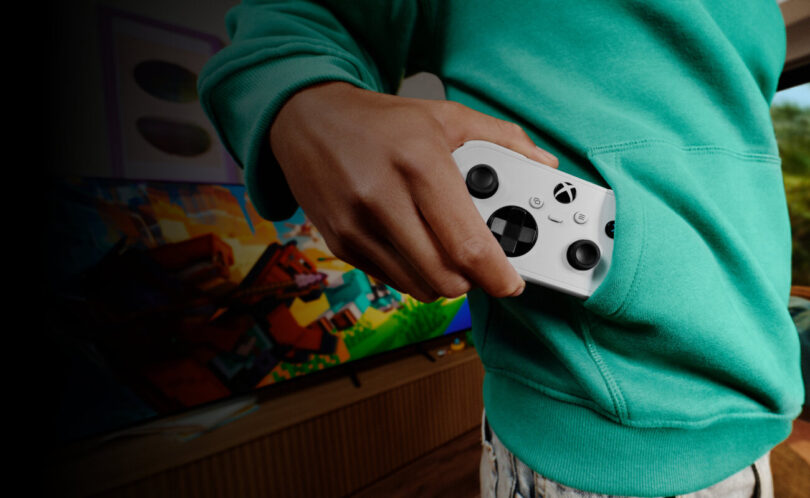 Manette XBOX Nano blanche glissée dans la poche d'un sweat vert, devant une TV affichant un jeu, illustrant le format de poche de la nouvelle manette Xbox pour le cloud gaming