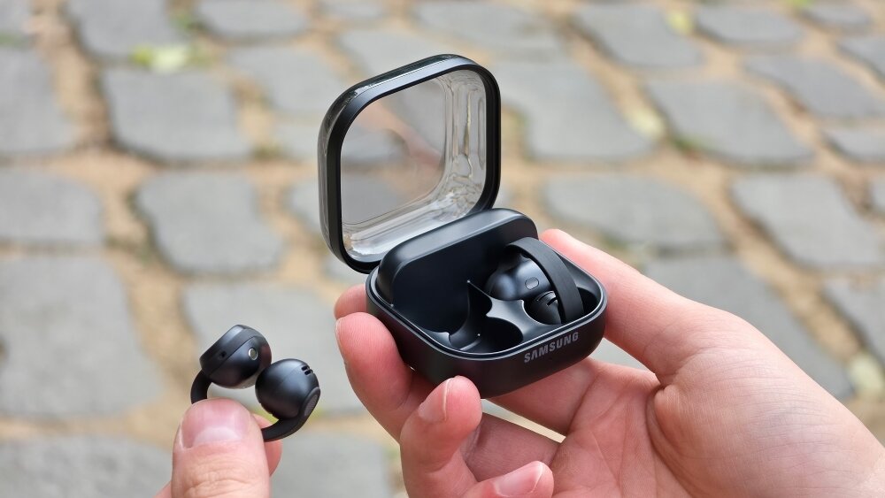 Main tenant le boîtier de charge ouvert des Samsung Galaxy Buds On noirs, un écouteur à clip sorti entre les doigts