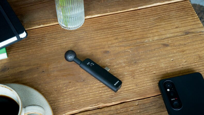 Le micro Sony ECM-AX10 posé sur une table en bois à côté d'un smartphone, avec son module sphérique à quatre capsules et sa prise USB-C