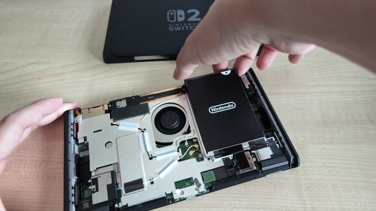 Batterie de la Nintendo Switch 2 révisée BEE-001(-01) soulevée hors de la console ouverte, à côté du ventilateur et de la carte mère