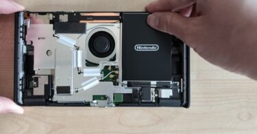 Intérieur de la Nintendo Switch 2 révisée BEE-001(-01) ouverte, avec le ventilateur, les caloducs, la carte mère et la batterie noire Nintendo que l'on soulève sans adhésif