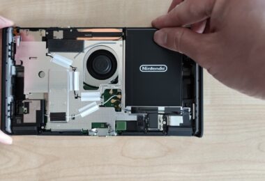 Intérieur de la Nintendo Switch 2 révisée BEE-001(-01) ouverte, avec le ventilateur, les caloducs, la carte mère et la batterie noire Nintendo que l'on soulève sans adhésif