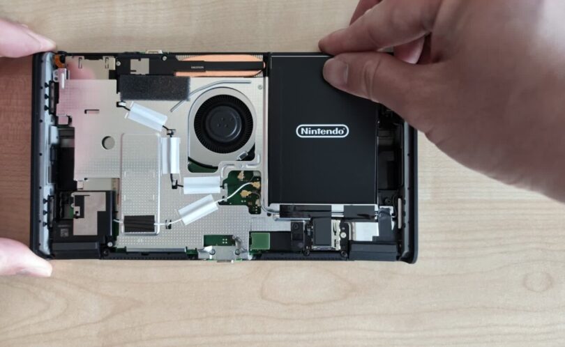 Intérieur de la Nintendo Switch 2 révisée BEE-001(-01) ouverte, avec le ventilateur, les caloducs, la carte mère et la batterie noire Nintendo que l'on soulève sans adhésif