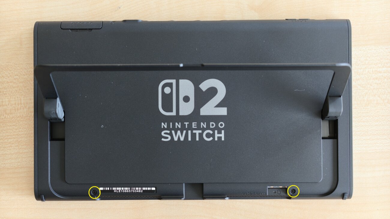 Dos de la Nintendo Switch 2 à batterie remplaçable avec le pied déplié, deux vis cruciformes entourées en jaune en bas de la coque arrière par le guide officiel Nintendo