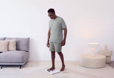 Homme se pesant pieds nus sur une balance connectée Withings Body Smart blanche dans un salon lumineux, illustration de la synchronisation des données avec Apple Santé