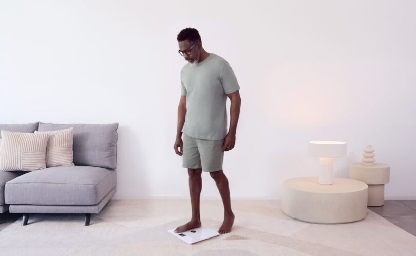 Homme se pesant pieds nus sur une balance connectée Withings Body Smart blanche dans un salon lumineux, illustration de la synchronisation des données avec Apple Santé