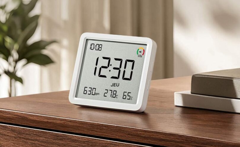 Le Xiaomi Smart Temperature and Humidity Monitor 3 Pro posé sur un meuble en bois, écran LCD affichant l'heure, le taux de CO2 en ppm, la température et l'humidité